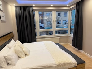 A bedroom with a large bed, a window with blinds, a radiator, and a view of buildings outside.