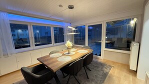 Un espace repas avec une table en bois, des chaises noires, un vase et une vue dégagée sur l’extérieur grâce à de grandes fenêtres.
