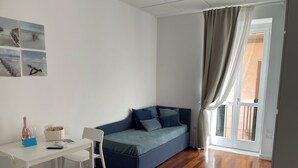 Une pièce avec un plancher en bois, un bureau blanc avec une chaise, un canapé bleu avec des coussins et une fenêtre avec des rideaux.