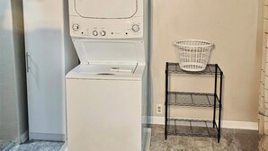 A laundry room with a stacked washer and dryer, a small metal shelf with a basket, and an air conditioning unit on the wall. - Cozy 1-bedroom apartment in tranquil Huntington with AC (Huntington)