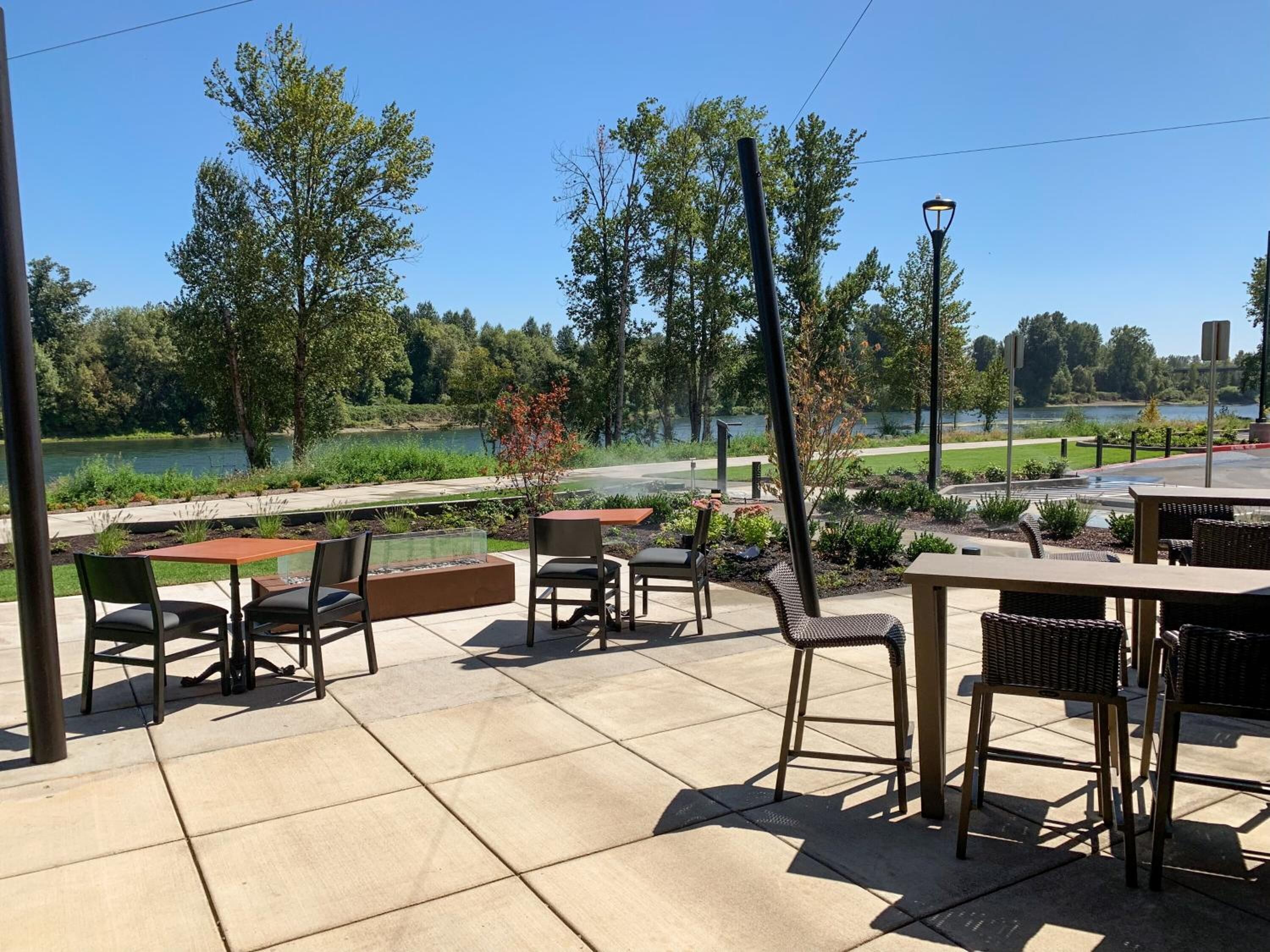 An outdoor patio with tables and chairs, a fire pit, and a view of a lake and trees.
