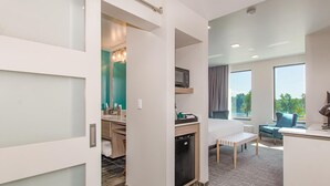 A hotel room with a bed, a desk, a microwave, a small refrigerator, a window with a view, and a chair.
