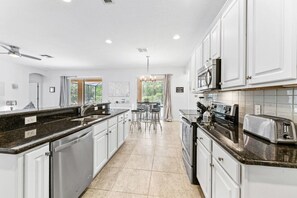 A modern kitchen with white cabinets, granite countertops, stainless steel appliances, and a dining area with bar stools. - Luxurious 5 bed Solterra Oasis close to Disney (Davenport)