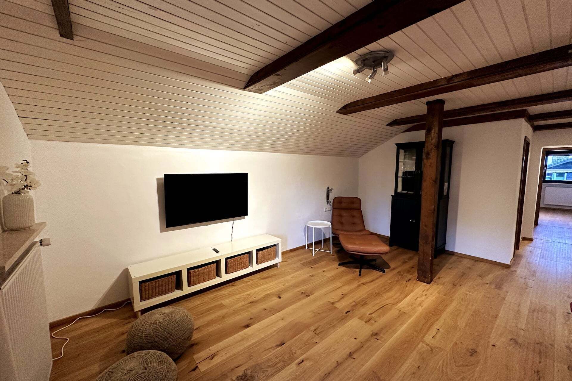 Ein Zimmer mit einem an der Wand befestigten Flachbildfernseher, einem braunen Ledersessel, einem kleinen weißen Beistelltisch und Holzboden.