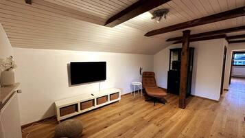 Ein Zimmer mit einem an der Wand befestigten Flachbildfernseher, einem braunen Ledersessel, einem kleinen weißen Beistelltisch und Holzboden.