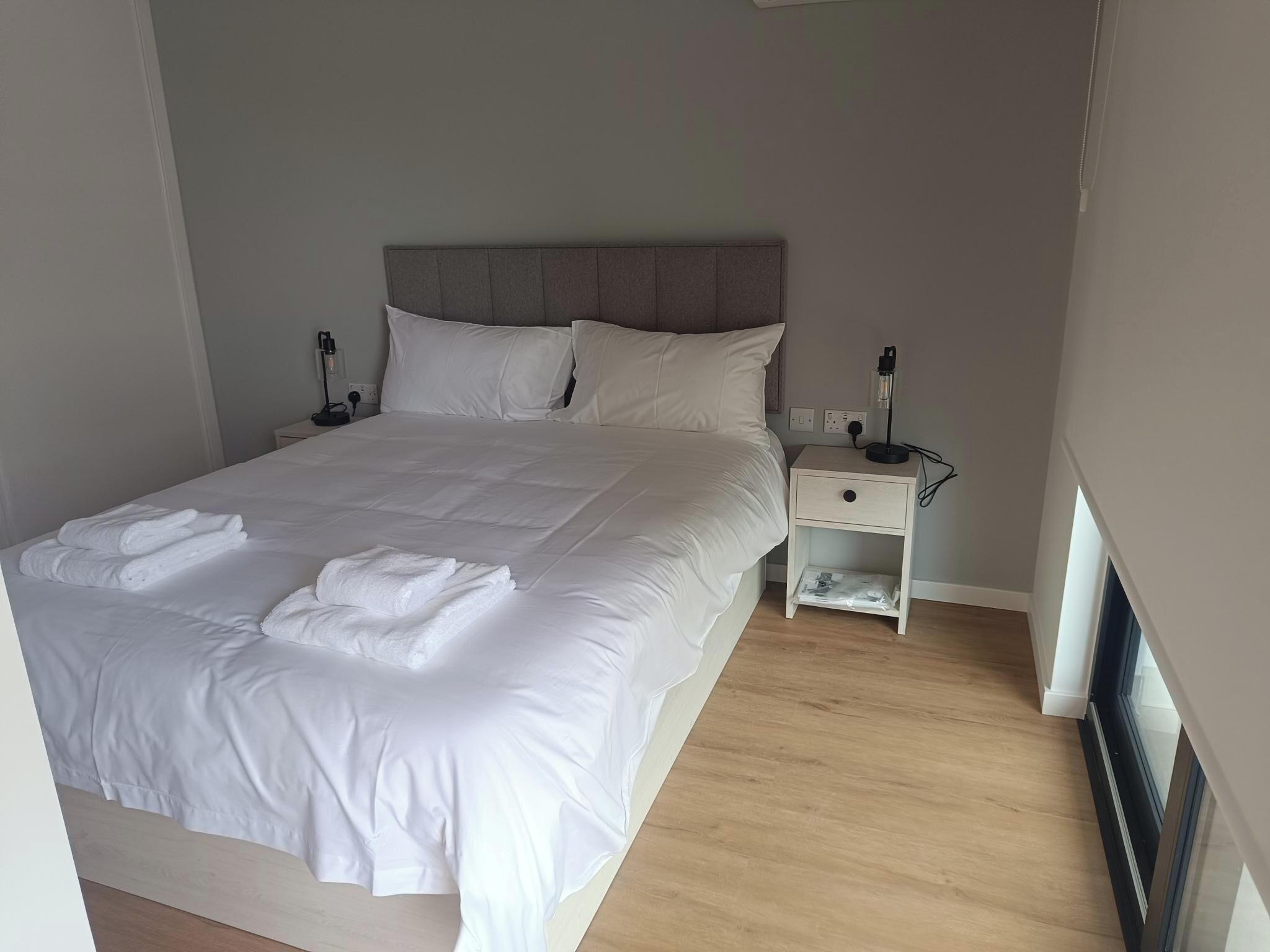A neatly made bed with white linens, a bedside table with a lamp, and a small drawer unit.