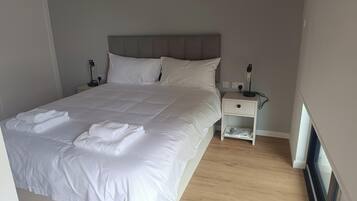 A neatly made bed with white linens, a bedside table with a lamp, and a small drawer unit.