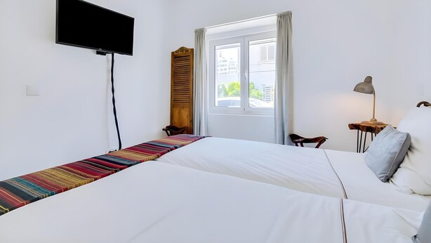 Quarto de hotel com cama, televisão na parede, janela com cortinas, mesinha com abajur e uma cadeira.