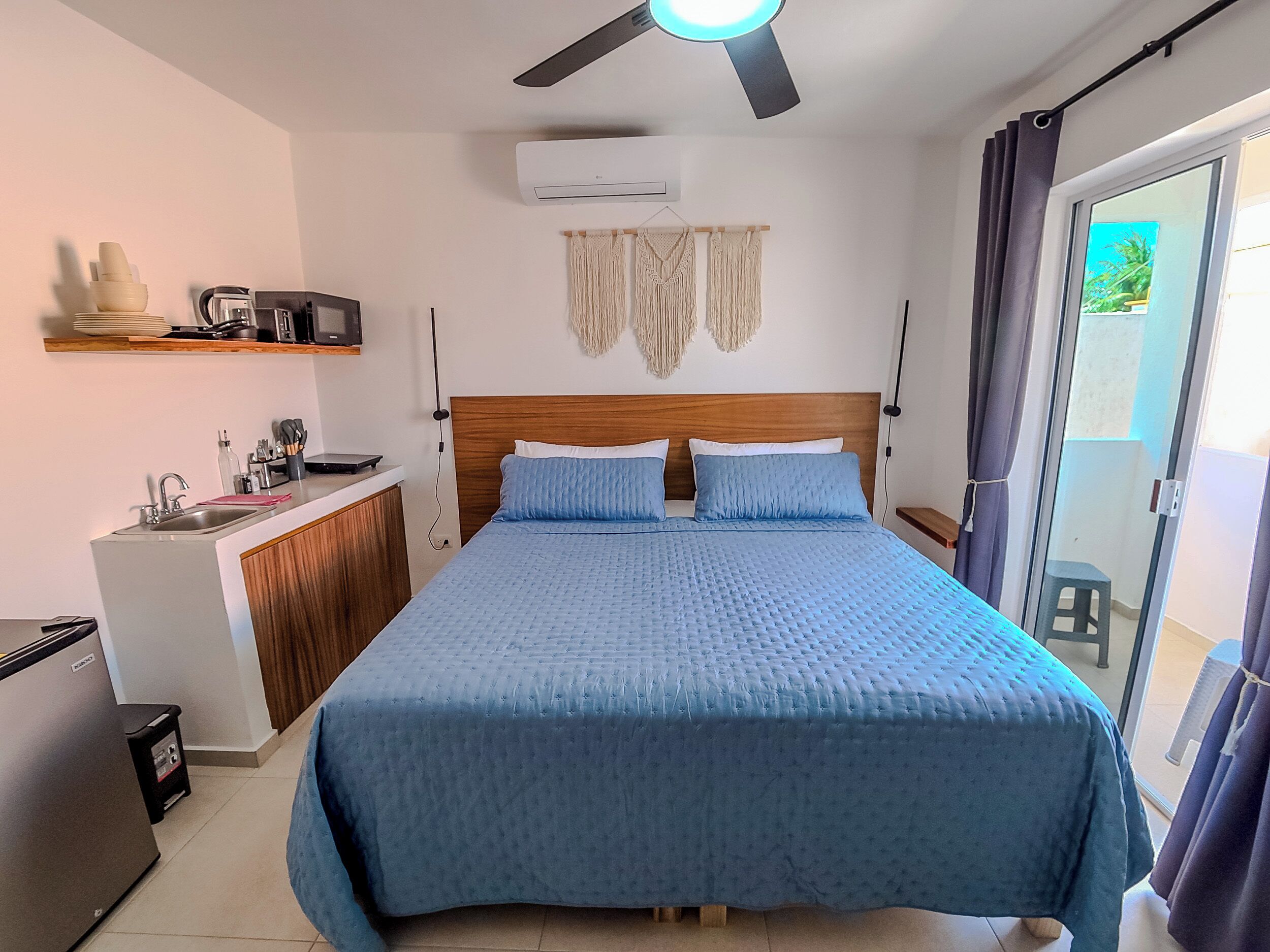 A bedroom with a bed, a ceiling fan, an air conditioner, a kitchenette, and a sliding glass door to the outside.
