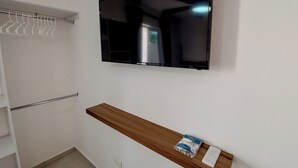 Ein Zimmer mit einem an der Wand befestigten Fernseher, einem Holzboden mit einer Fernbedienung und einem weißen Kleiderschrank mit Kleidung.