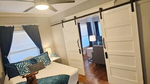 Quarto com cama, ventoinha no teto, janela grande com cortinas, porta deslizante e vista para outro cômodo.