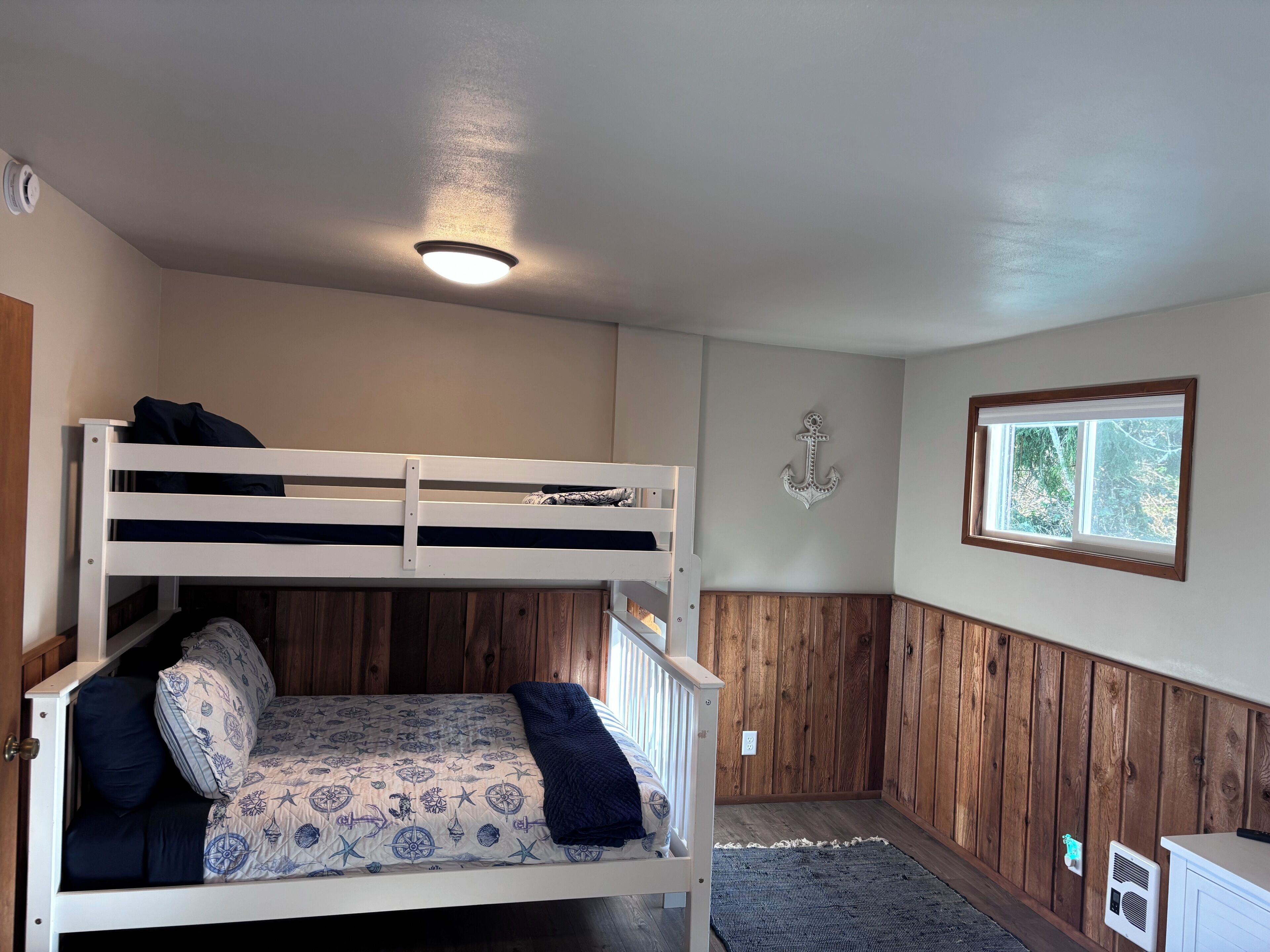A bunk bed with a patterned bedspread, a ceiling light, a wooden wall panel, a decorative anchor, and a window with a view.