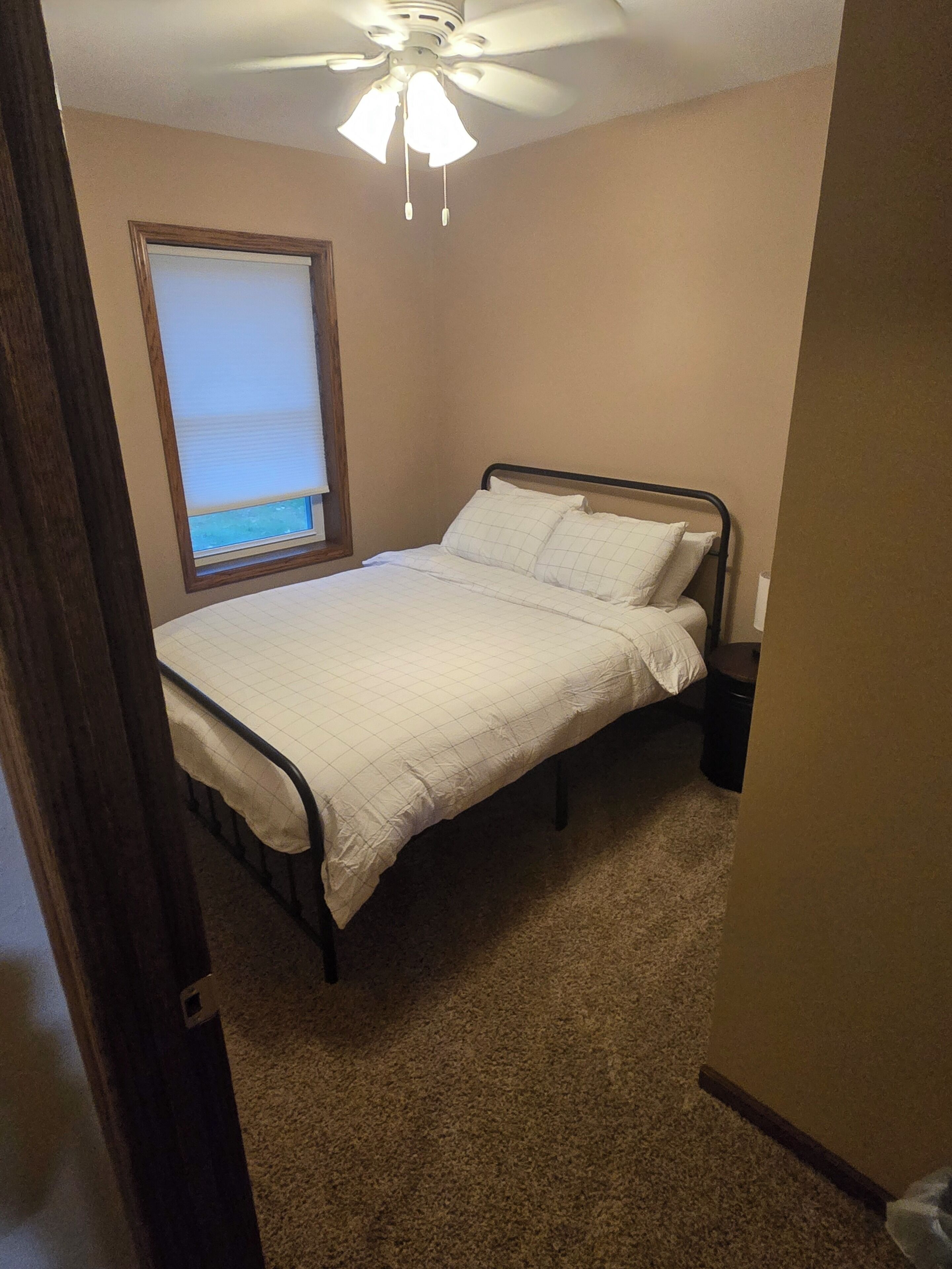 Een slaapkamer met een bed, een plafondventilator, een raam met zonwering en een vloerbedekking.