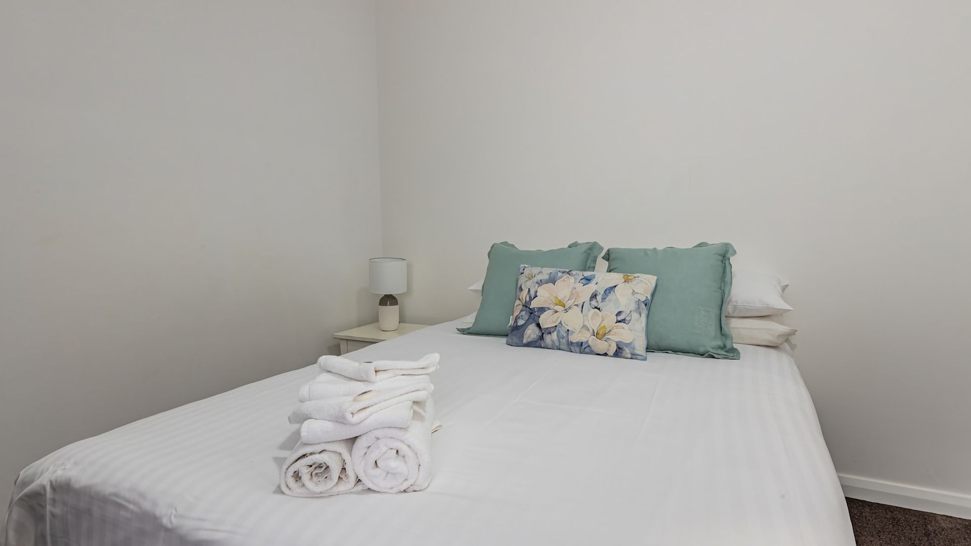 A neatly made bed with white linens and rolled towels, a floral pillow, and two teal pillows on a plain white wall.