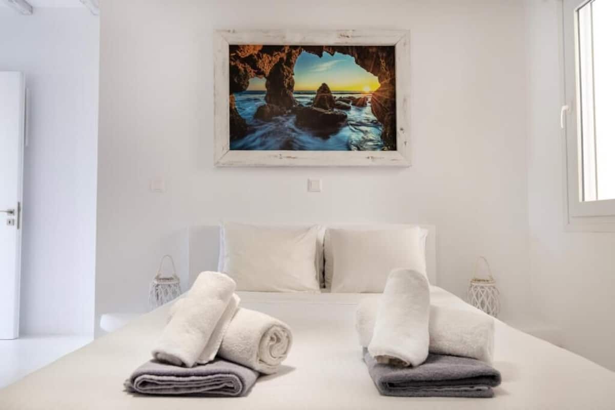 Habitación de hotel con cama, dos toallas enrolladas, un cuadro con un atardecer sobre rocas y una puerta blanca.