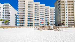 Un hotel frente a la playa con varios balcones, una playa arenosa en primer plano y un paseo de madera que conduce a la entrada.