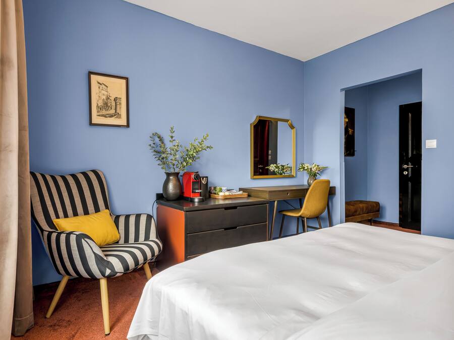 Ein Hotelzimmer mit einem Bett, einem Schreibtisch, einem Sessel, einer Kommode, einem Spiegel und einem gerahmten Bild an der Wand.