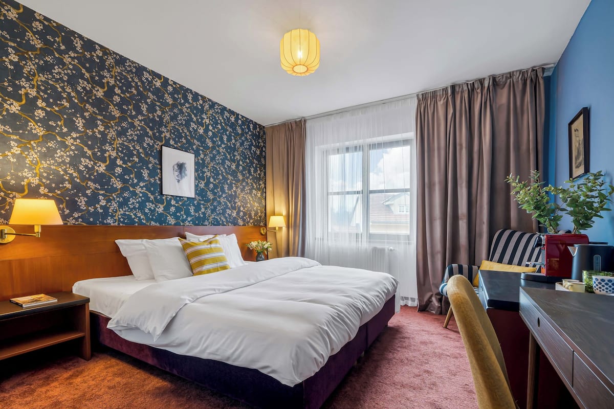 A hotel room with a large bed, a desk with a chair, a lamp, and a window with curtains.