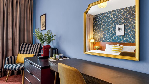 A hotel room with a bed, a desk with a chair, a dresser, a coffee maker, a plant, and a large mirror. - Penzion V Holi (Průhonice)