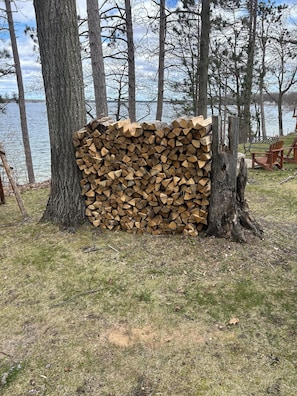 Ein Holzstapel an einem Baum, ein See und Stühle im Hintergrund.
