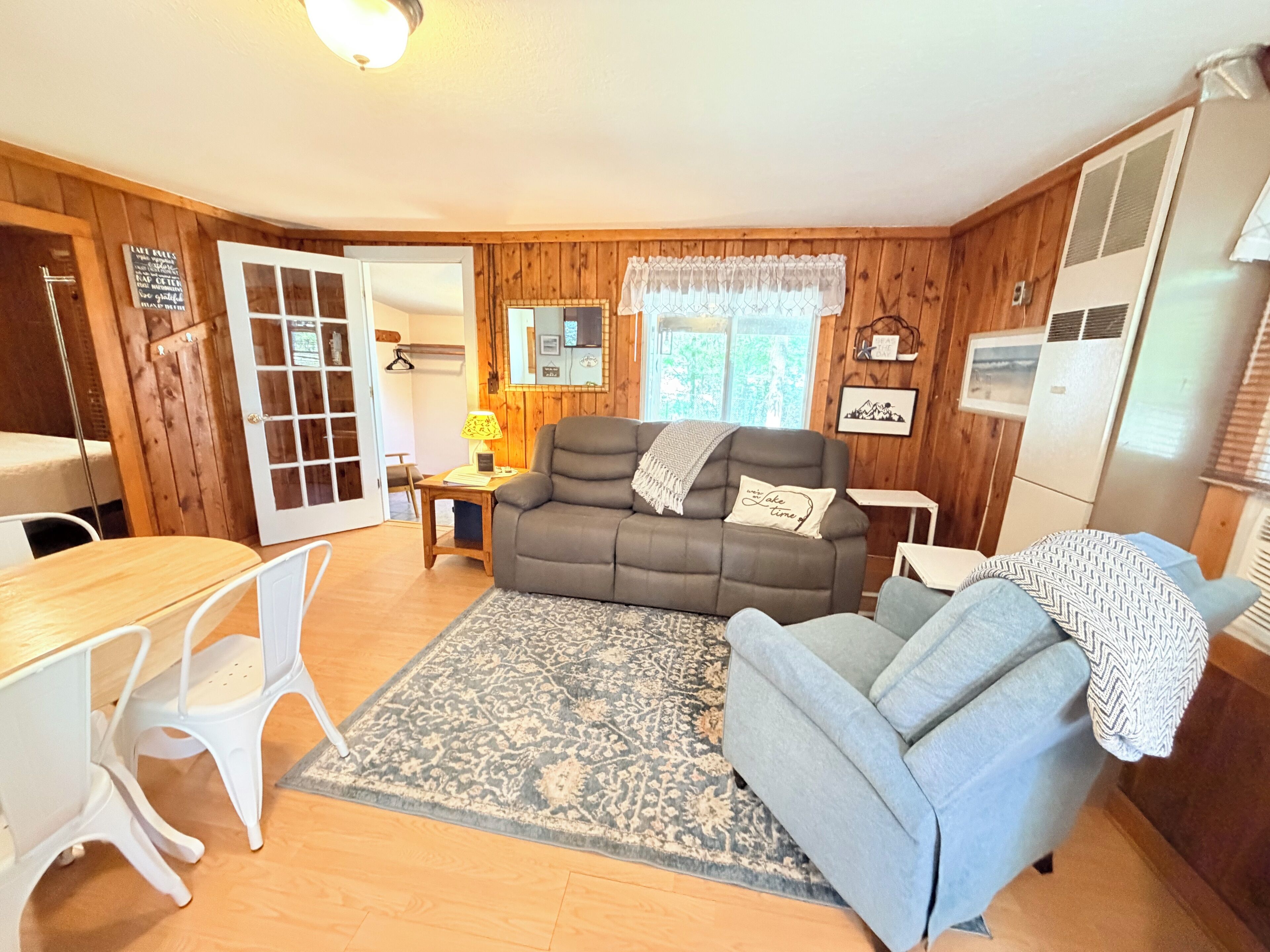 A cozy room with a sofa, a dining table, and chairs, wooden walls, and a window with a view.
