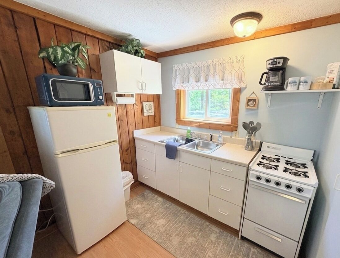 A compact kitchen with a refrigerator, microwave, sink, stove, and wooden walls.