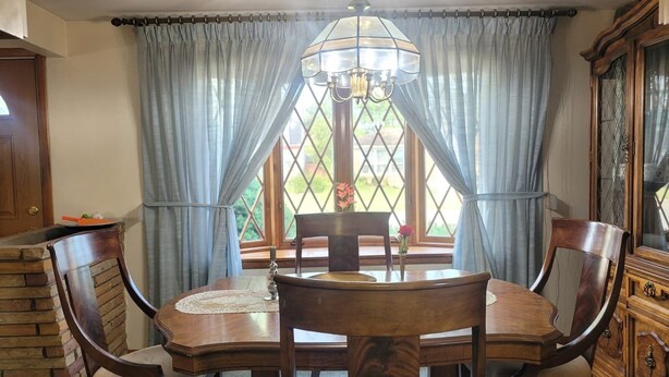 Une salle à manger avec une table ronde en bois, quatre chaises, un lustre et une fenêtre avec des rideaux.