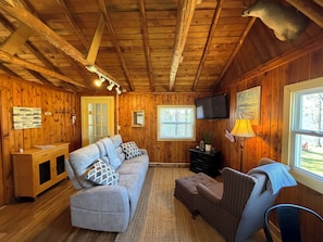 A cabin interior with wooden walls, a sofa, a television, and a window with a view.