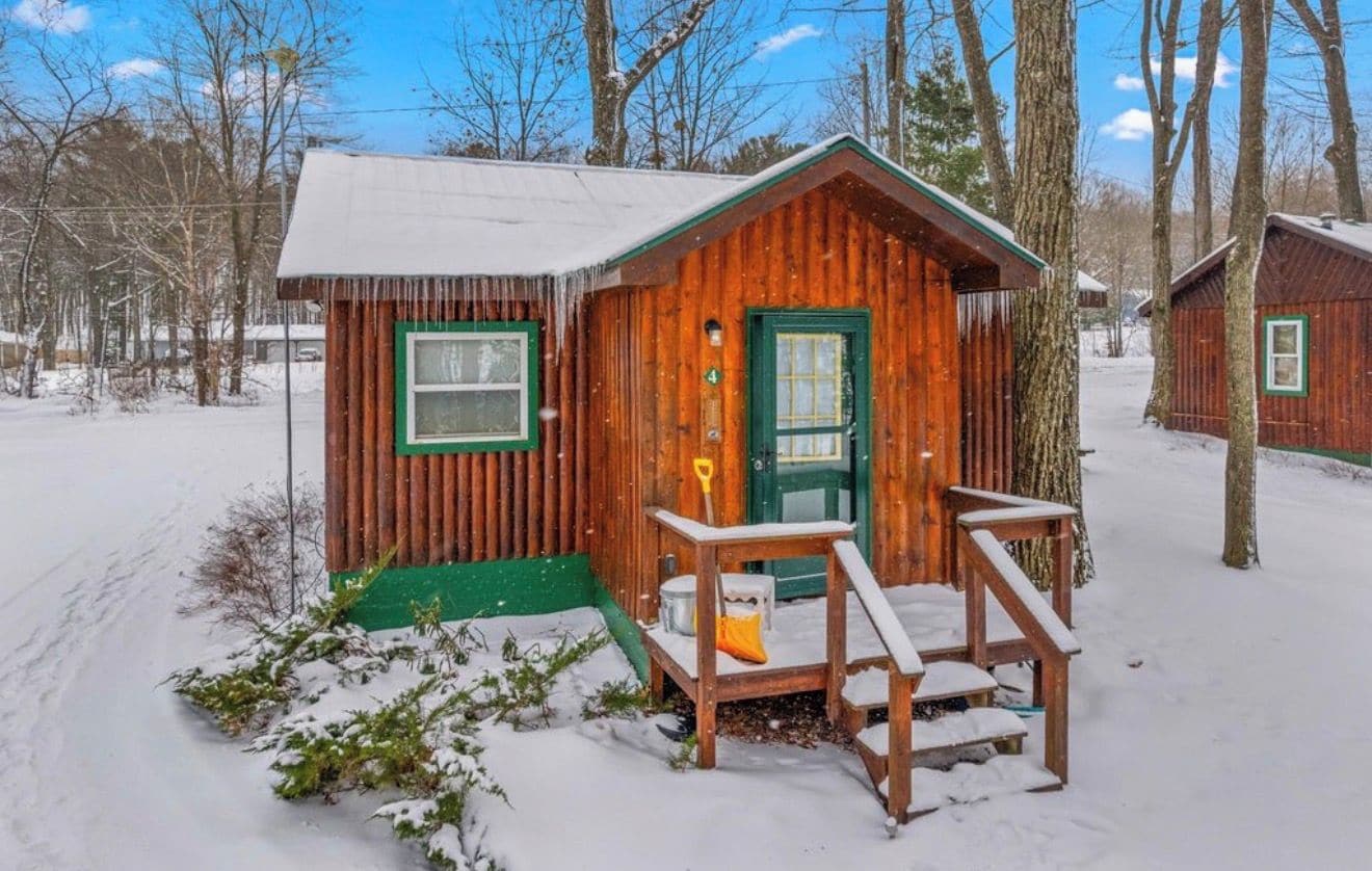 Une cabane en bois avec une porte et des fenêtres vertes, entourée d’un paysage enneigé et d’arbres.