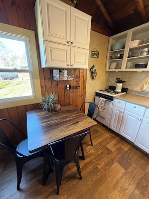 A wooden dining table with chairs, a kitchen with white cabinets, a stove, and a window with a view of the outdoors.