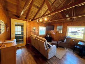 A cabin interior with a sofa, armchairs, a television, wooden walls, and a window view.