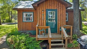 Una baita di legno con una porta verde, un piccolo portico con una panca e un barbecue esterno.