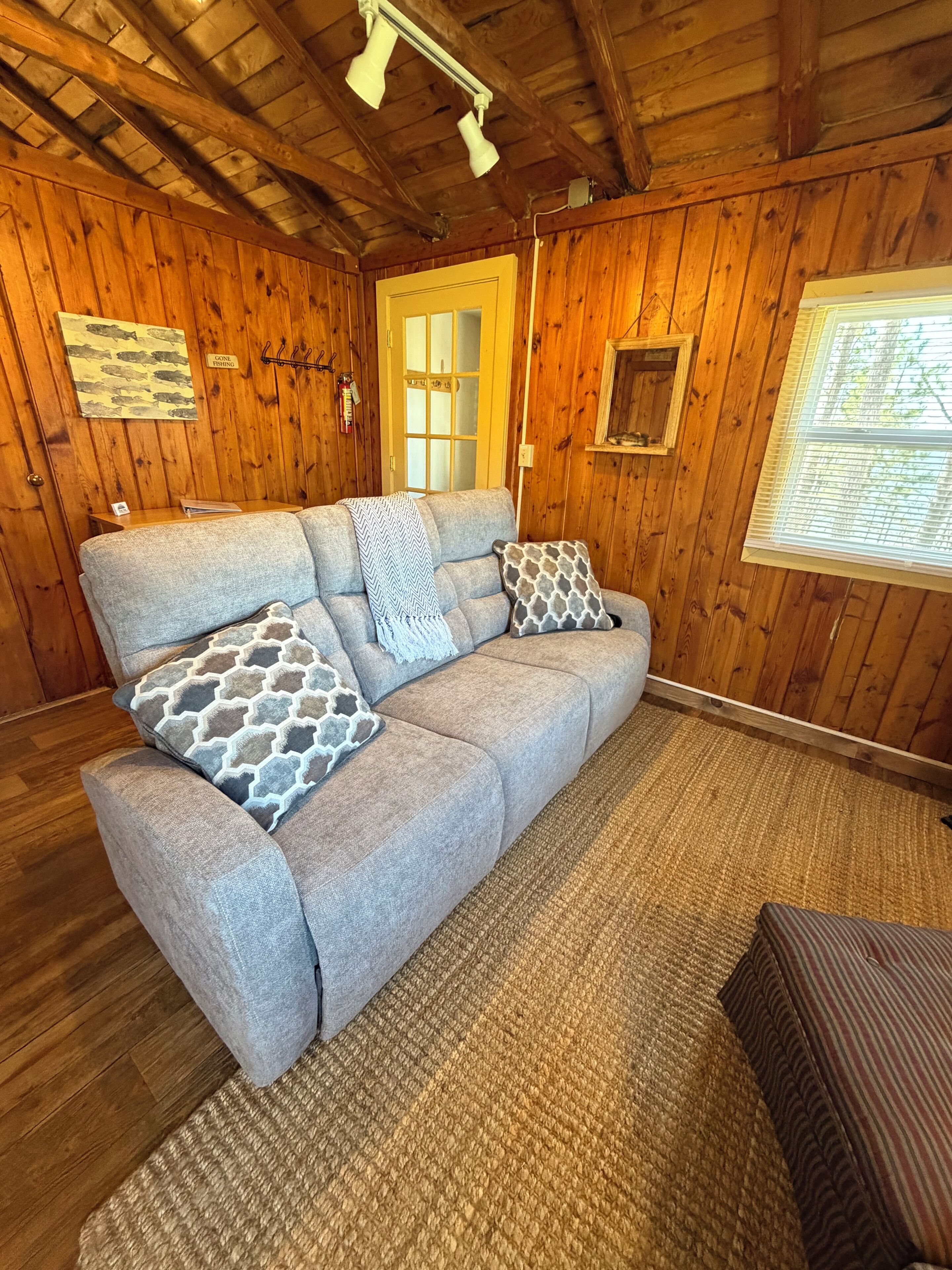 A wooden interior with a sofa, patterned cushions, a rug, and a window with blinds.