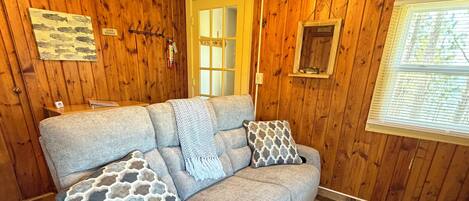 A wooden interior with a sofa, patterned cushions, a rug, and a window with blinds.