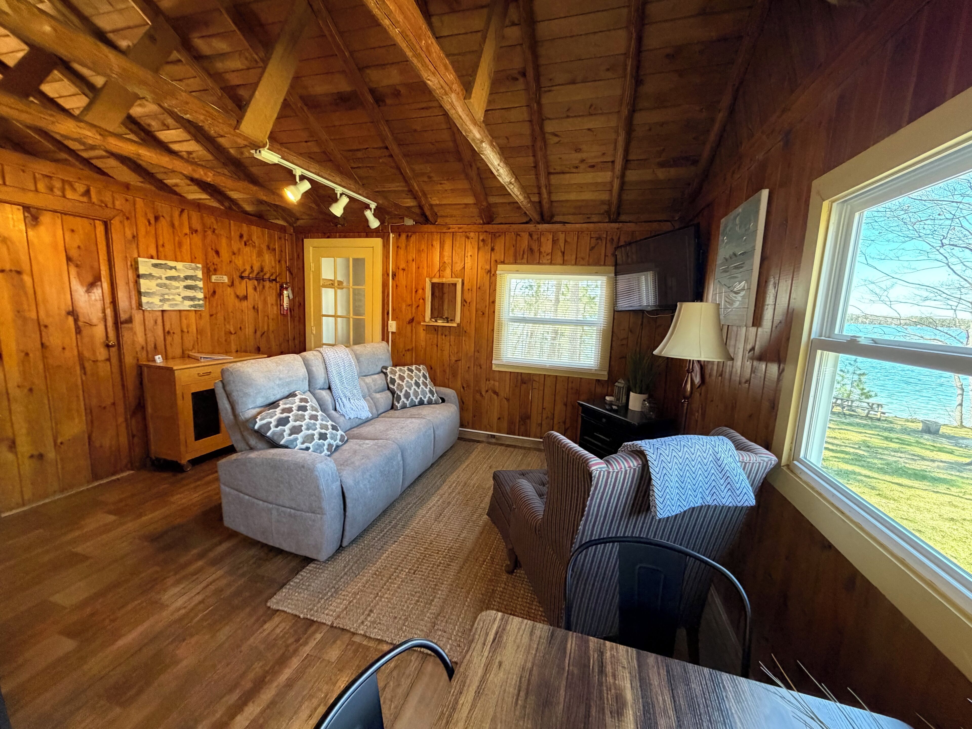 A cozy cabin interior with wooden walls, a sofa, a small table, and a window with a view of a lake.