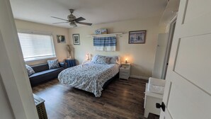 Une chambre à coucher comprenant un lit, des tables de chevet, un ventilateur de plafond, une fenêtre avec des stores et une étagère fixée au mur.
