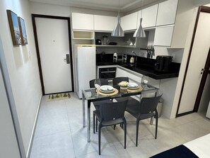 A compact kitchen with white cabinets, a black countertop, and a glass dining table with chairs. - Flat com piscina a 50m da praia (Recife)