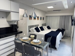 A modern hotel room with a bed, a dining area with a glass table, and a kitchenette. - Flat com piscina a 50m da praia (Recife)