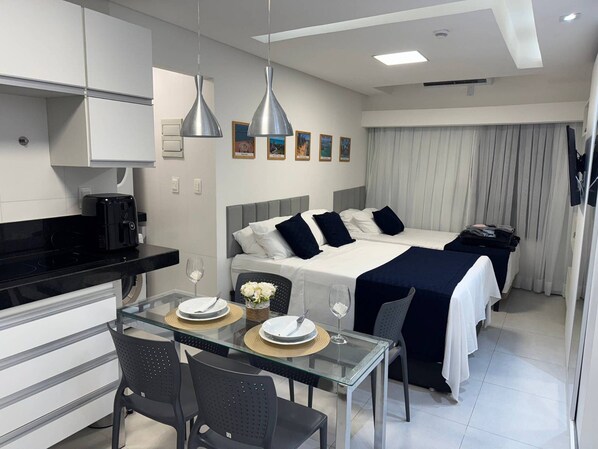 A modern hotel room with a bed, a dining area with a glass table, and a kitchenette.
