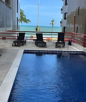 A swimming pool with lounge chairs, a view of the beach, and a tall structure in the background.