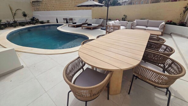Uma área à beira da piscina com mesa, cadeiras e espreguiçadeiras, além de um pequeno guarda-sol.