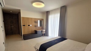 Quarto de hotel moderno com uma cama grande, uma escrivaninha, área de chuveiro e uma janela ampla com cortinas.