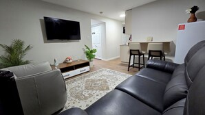 A living room with a sofa, a TV, a coffee table, a plant, and a kitchen area with a counter and chairs. - Stylish New 2BR Suite | Private Entry | Quiet, Near Shops & Transit (Winnipeg)