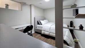 A compact room with a bed, a desk, and a chair. There is a window with blinds, a shelf with books, and a small decorative item on the shelf. - Stylish New 2BR Suite | Private Entry | Quiet, Near Shops & Transit (Winnipeg)