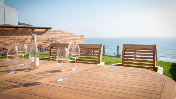 A wooden table with four chairs and wine glasses, overlooking a body of water. - Cornica - Pet Friendly Coastal House with Hot Tub and Sea Views (Portwrinkle)