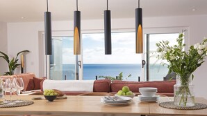 A dining area with a view of the sea, a sofa with a blanket, and a vase with flowers. - Cornica - Pet Friendly Coastal House with Hot Tub and Sea Views (Portwrinkle)