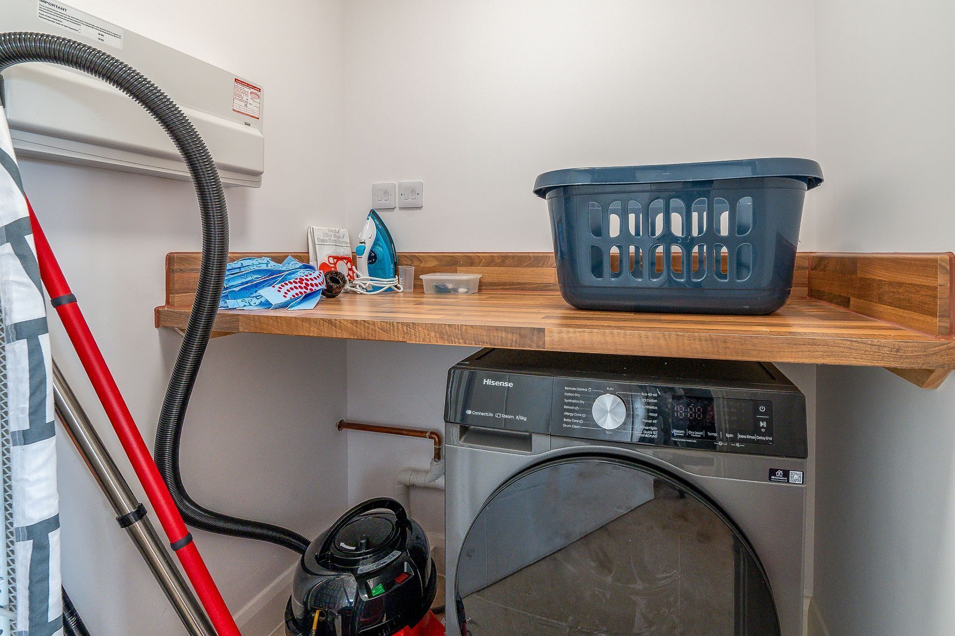 A laundry area with a washing machine, a vacuum cleaner, and a shelf with a laundry basket and cleaning supplies.