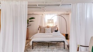 Une chambre à coucher avec un lit, des rideaux blancs, une lampe sur pied et une plante.
