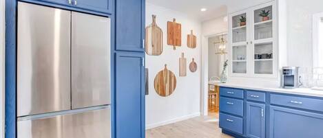 A modern kitchen with blue cabinets, a stainless steel refrigerator, wooden wall decorations, and a view into the dining area.