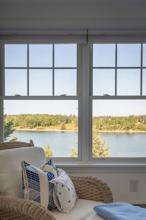 A room with a large window overlooking a lake, a wicker chair, and a cushion with a blue and white pattern. - Designer Waterfront Newly Renovated Cottage "ROXABOXEN" (Tremont)